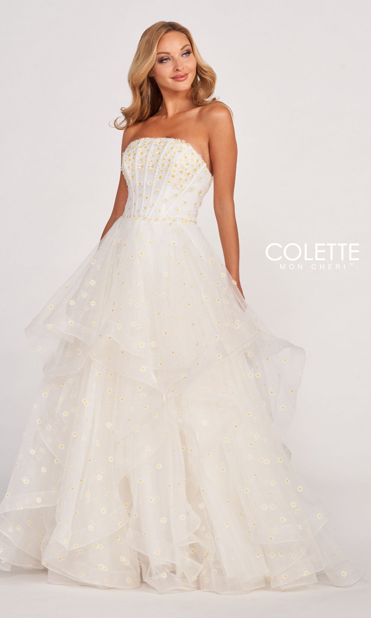 A woman stands against a plain light background, wearing the Colette CL2055 Strapless Daisy Ball Gown featuring a layered tulle skirt and gold floral accents, radiating elegance and timeless charm.